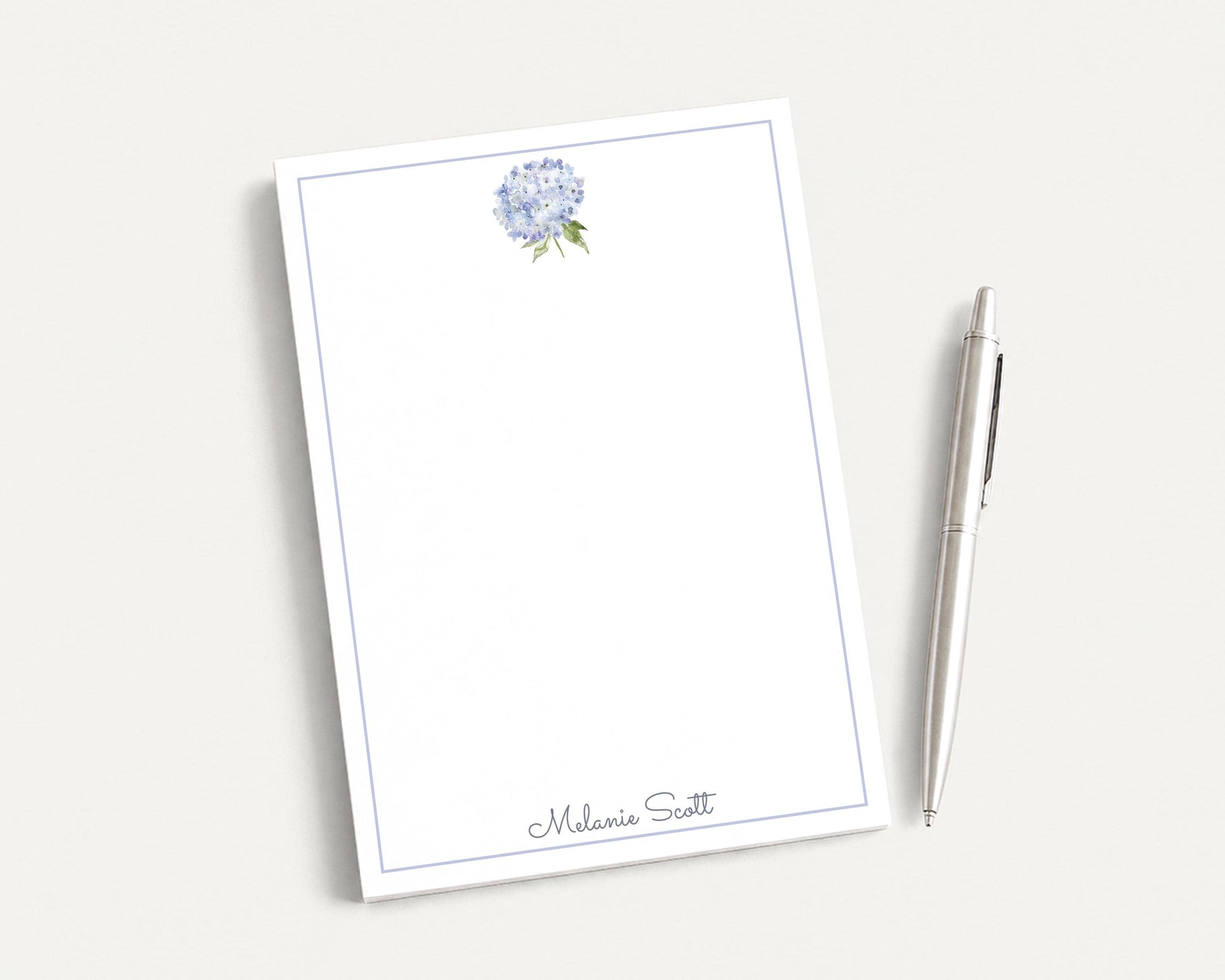 a blank white notepad with a blue border, and a silver pen is placed next to it on a white surface.
