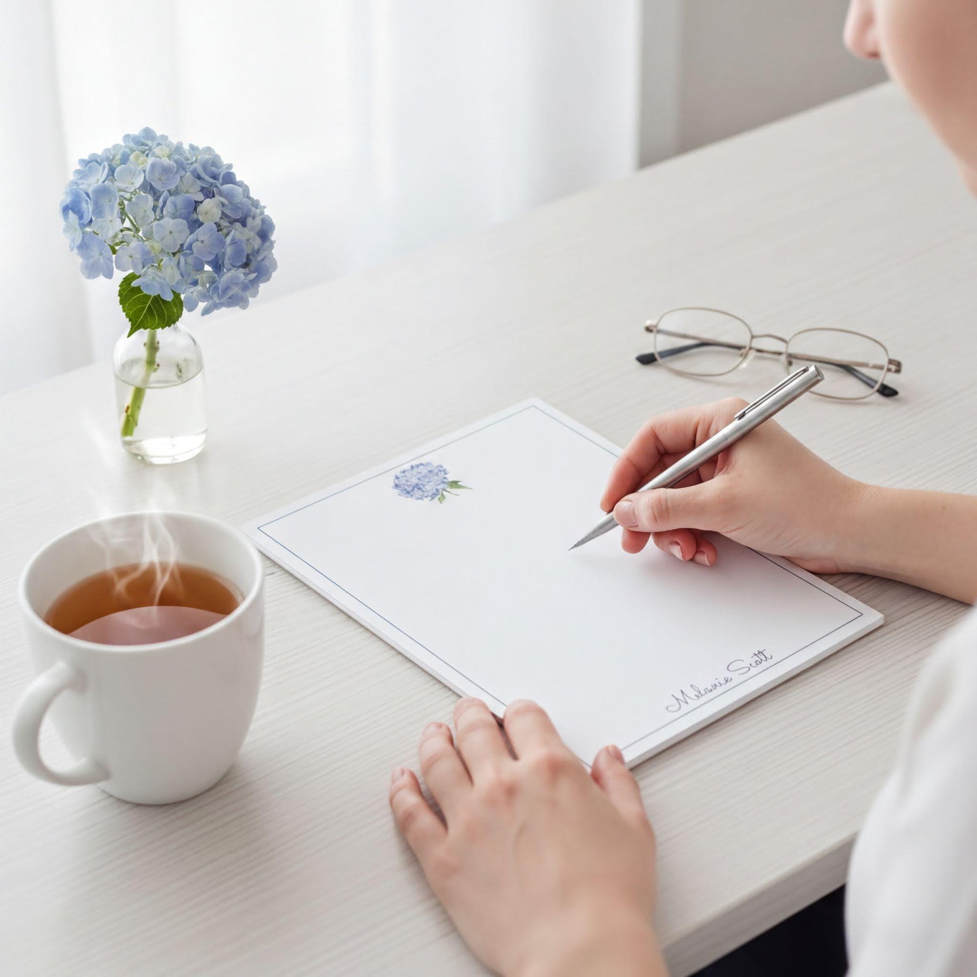 A person is sitting at a desk, writing on a piece of paper with a pen, while a cup of tea and a vase of blue hydrangeas are nearby.