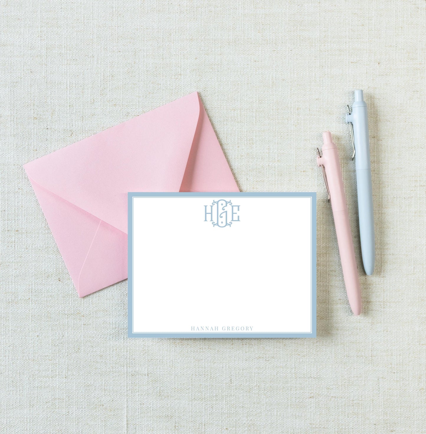 a pink envelope, a blank white card with a monogrammed logo, and two pens on a light-colored surface.