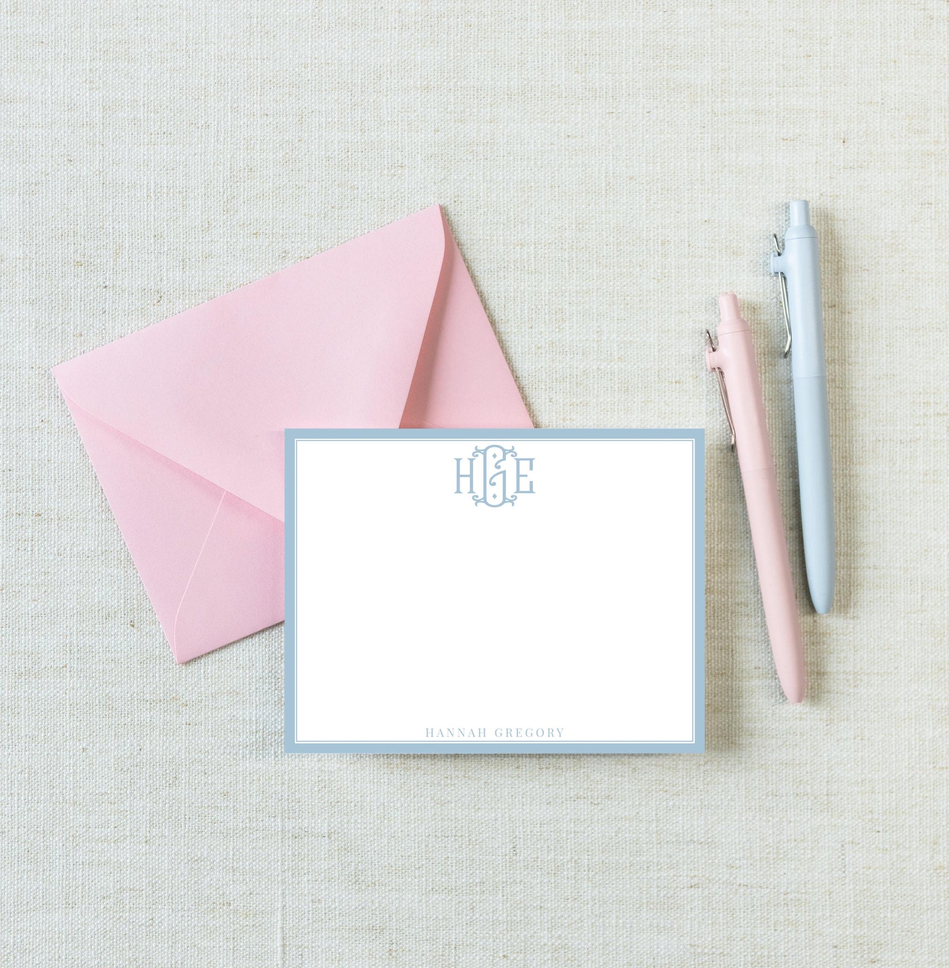 a pink envelope, a blank white card with a monogrammed logo, and two pens on a light-colored surface.