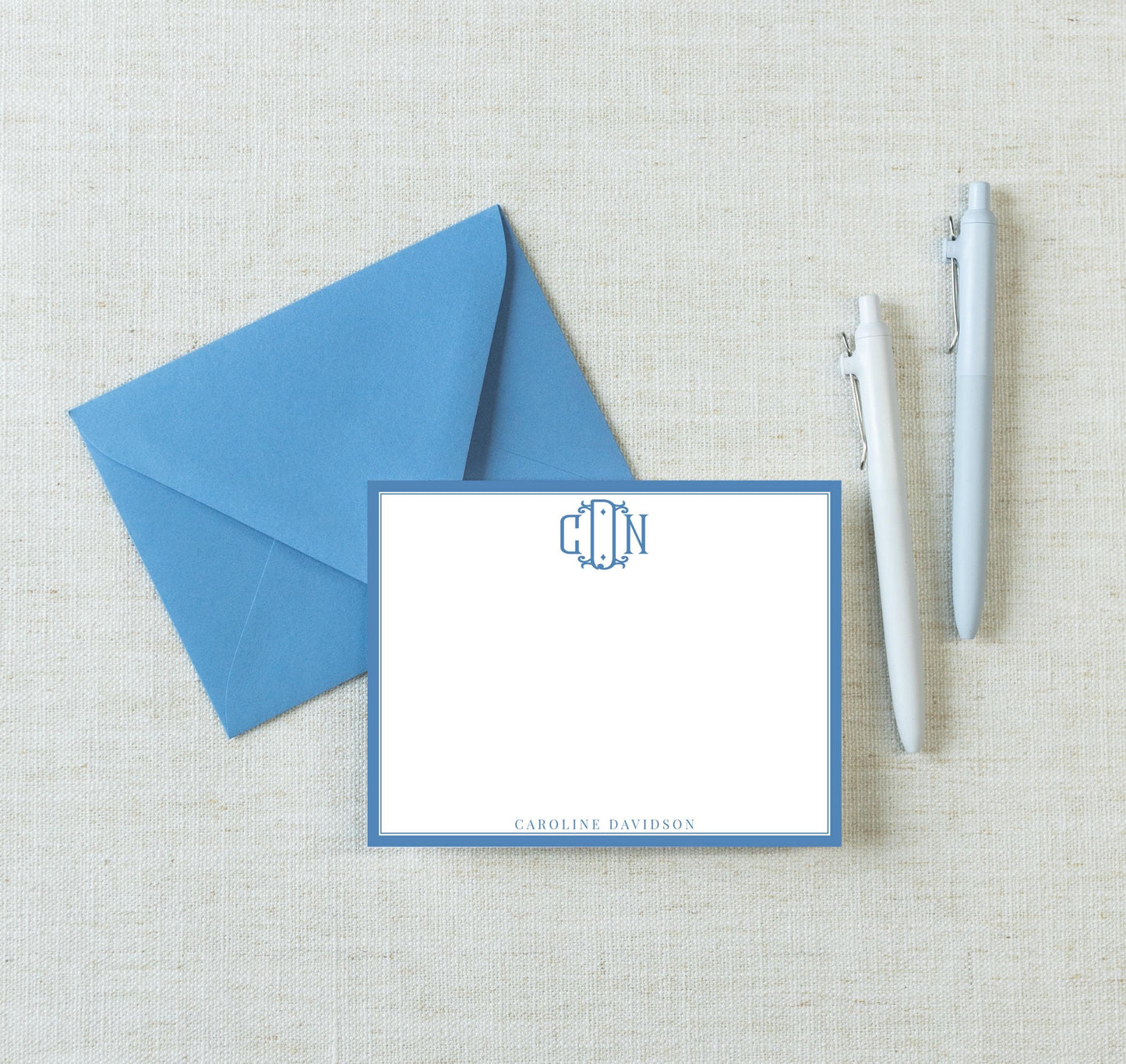 a blue envelope, a blank white card with a monogram, and two pens on a beige background.