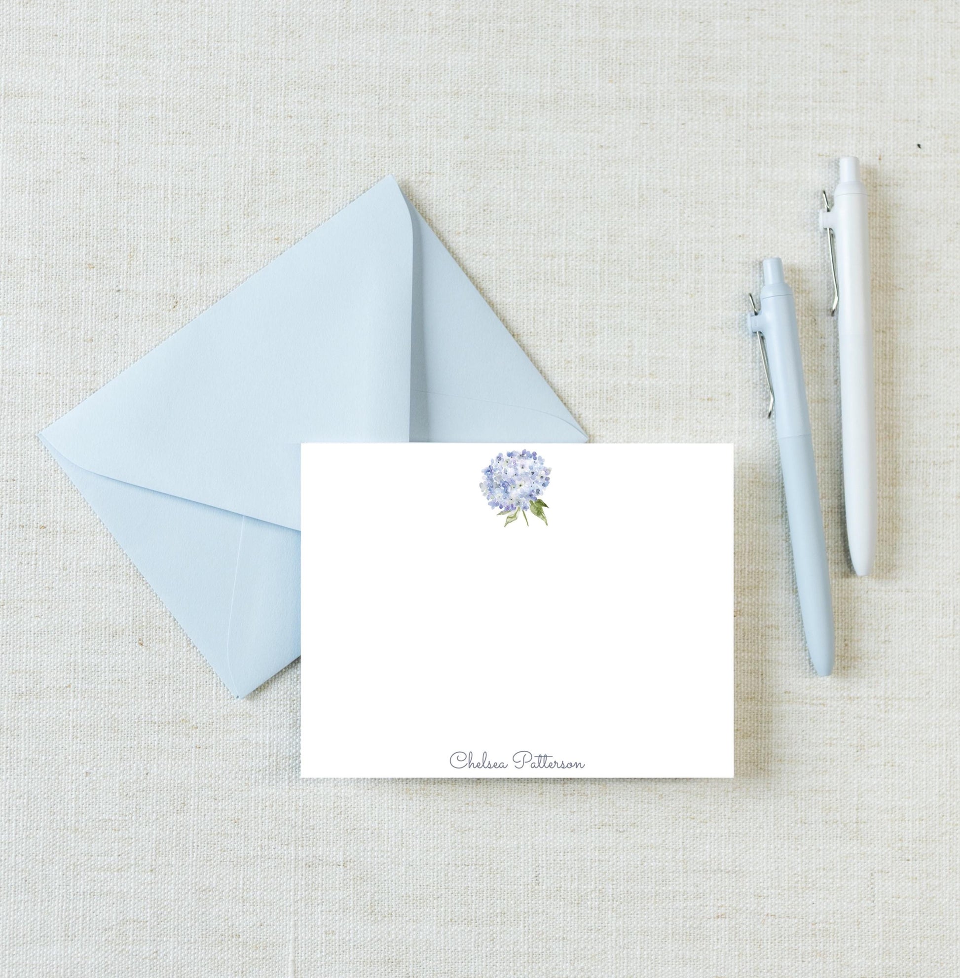 a light blue envelope with a white card inside, along with two pens and a light blue flower on a light beige background.