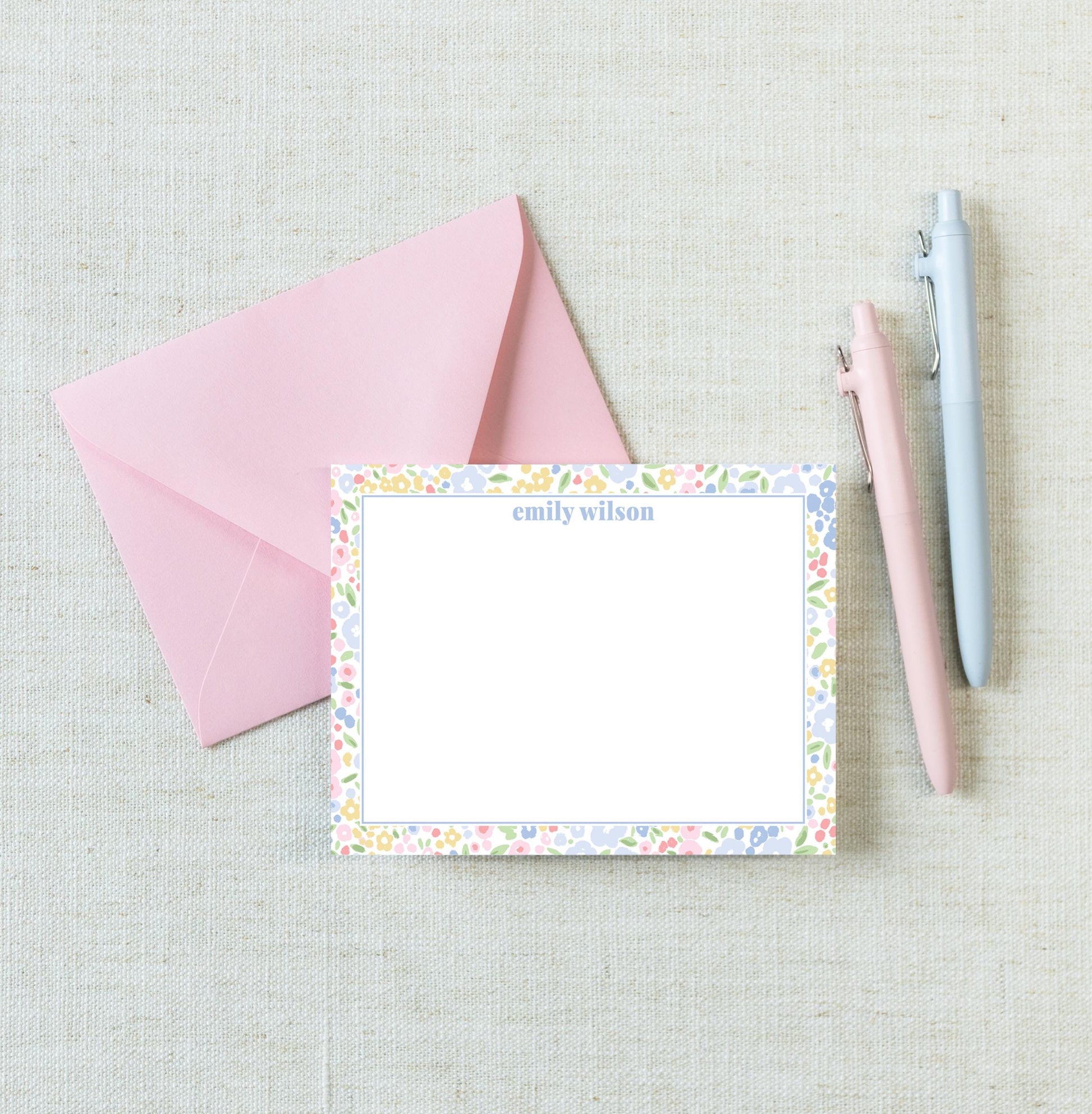 a pink envelope, a blank personalized note card with a floral border, and two pens on a light-colored surface.