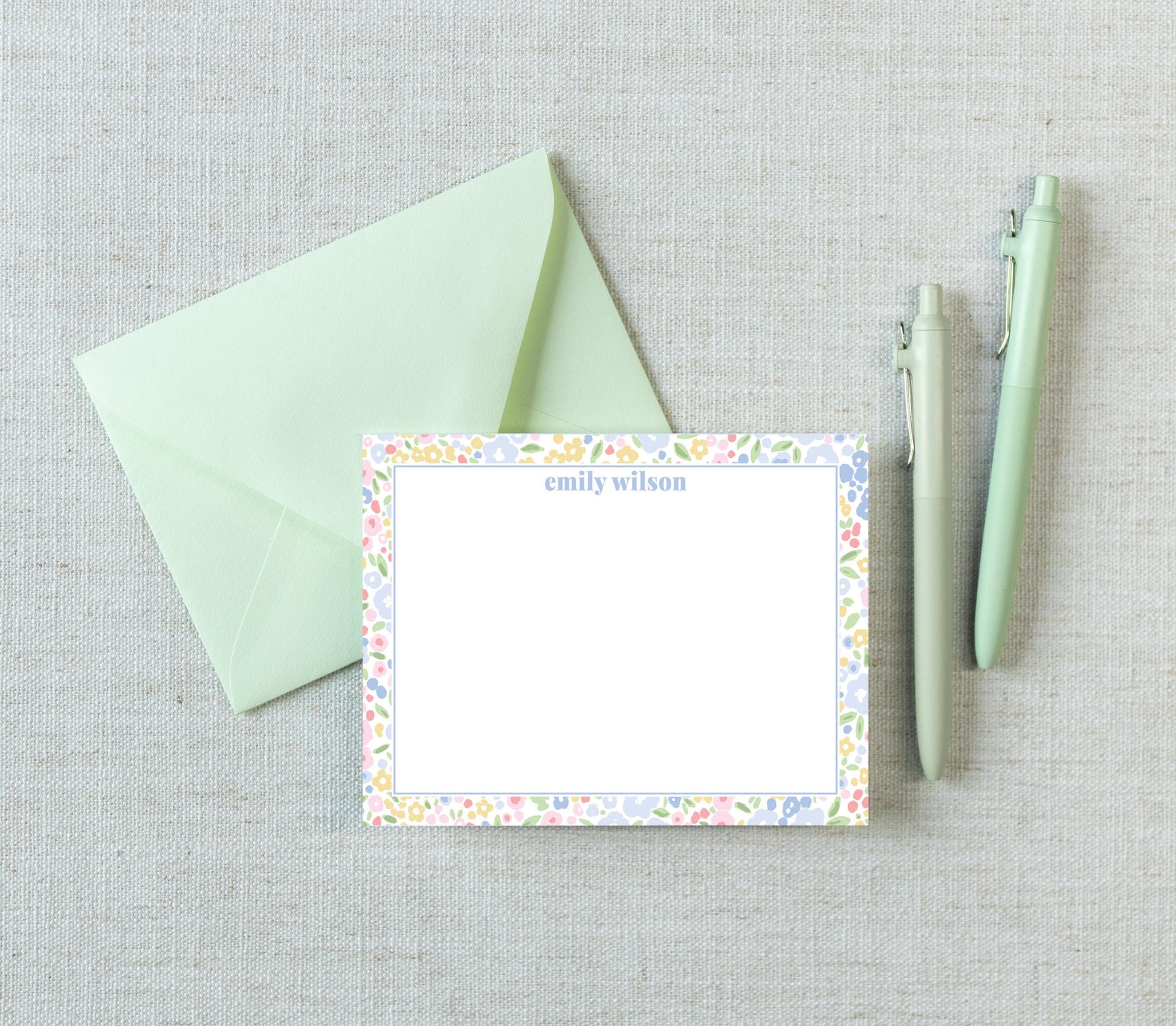 a green envelope, a blank card with a floral border, and two pens on a light-colored surface.