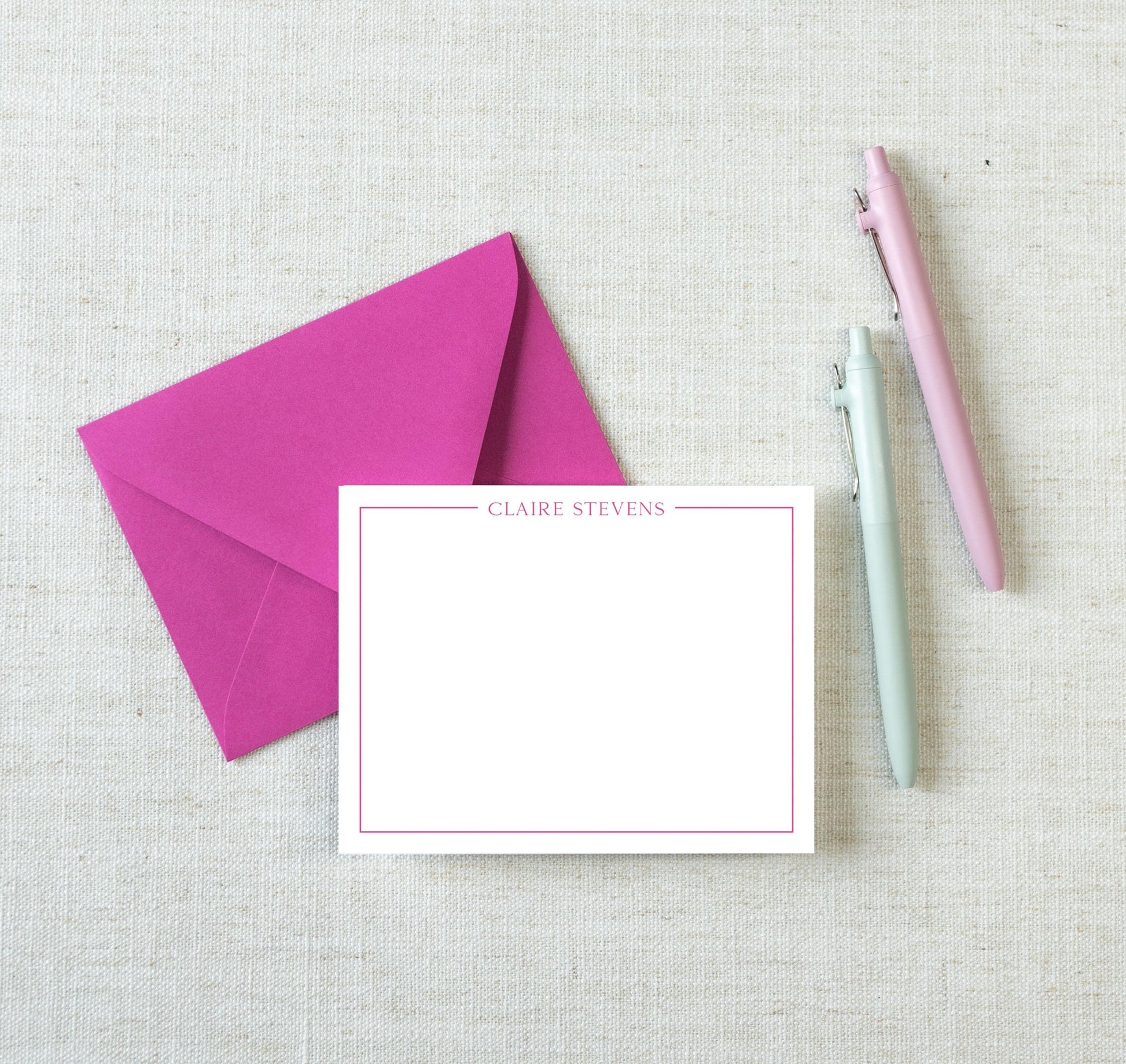 a pink envelope, a blank white card with a pink border, and two pens on a light-colored surface.