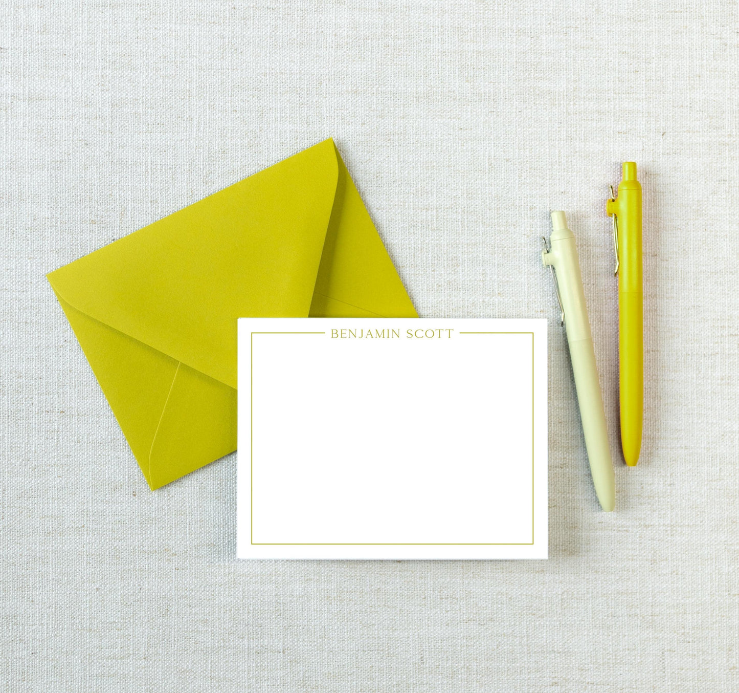 a yellow envelope, a blank white note card with the name "Benjamin Scott" written on it, and two yellow pens on a white background.