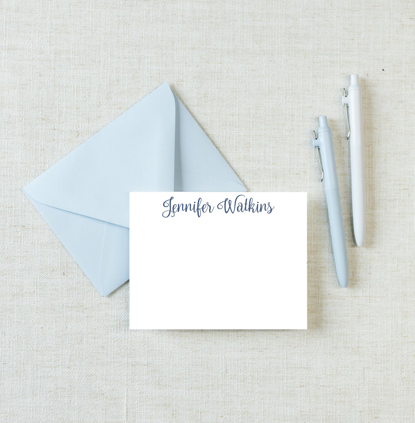 a light blue envelope with a blank white card inside, along with two pens on a light beige background.
