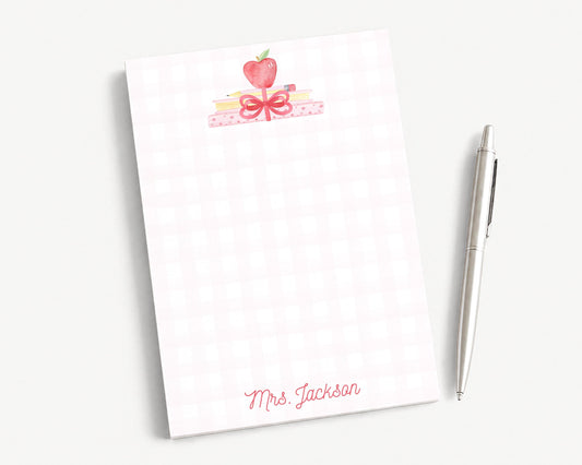 A white notepad with a pink and red design, featuring a heart and a bow, along with a pen and a pencil on a white surface.