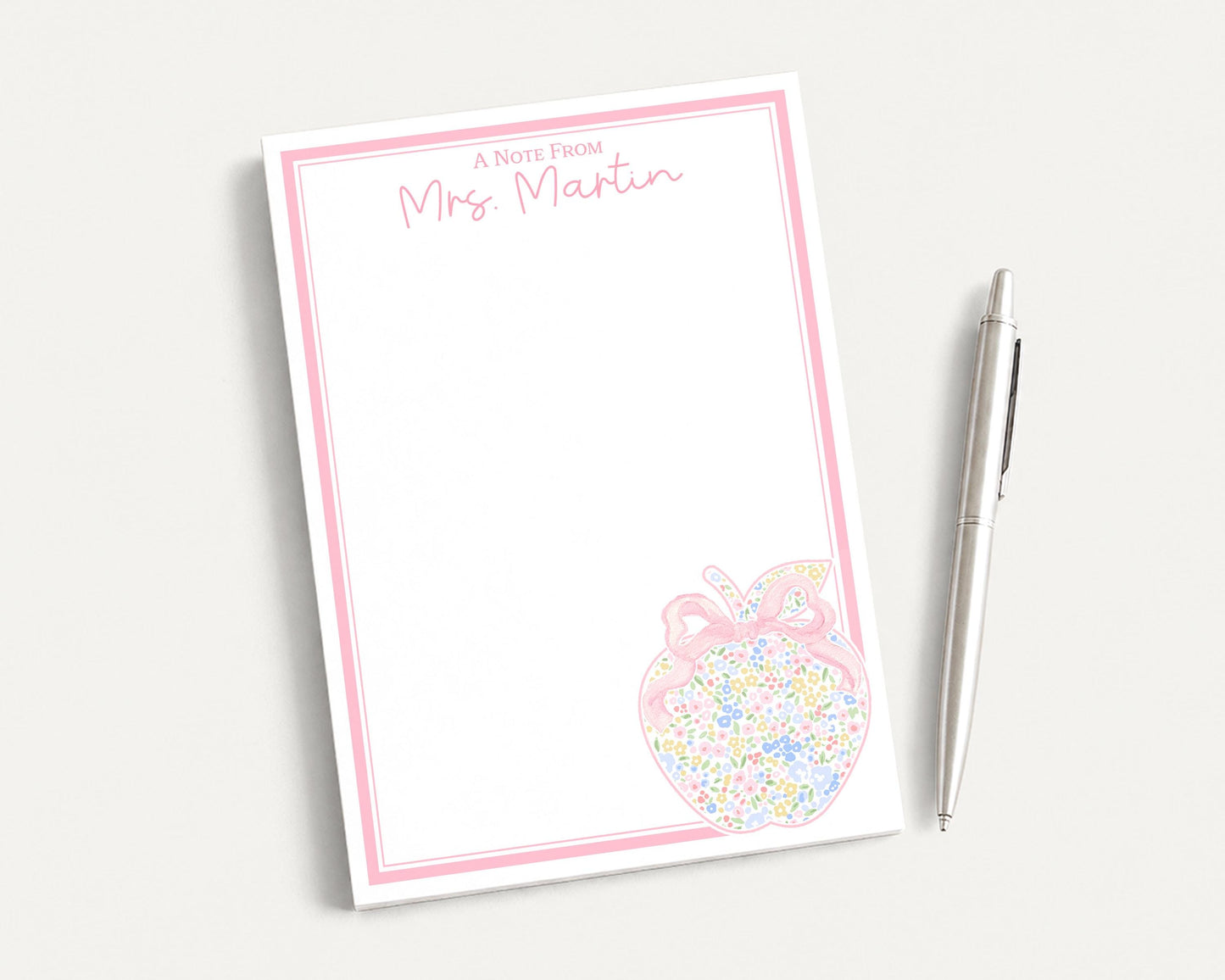 A blank note pad with a pink border and an illustration of an apple on it, accompanied by a pen on a white surface.
