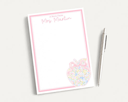A blank note pad with a pink border and an illustration of an apple on it, accompanied by a pen on a white surface.