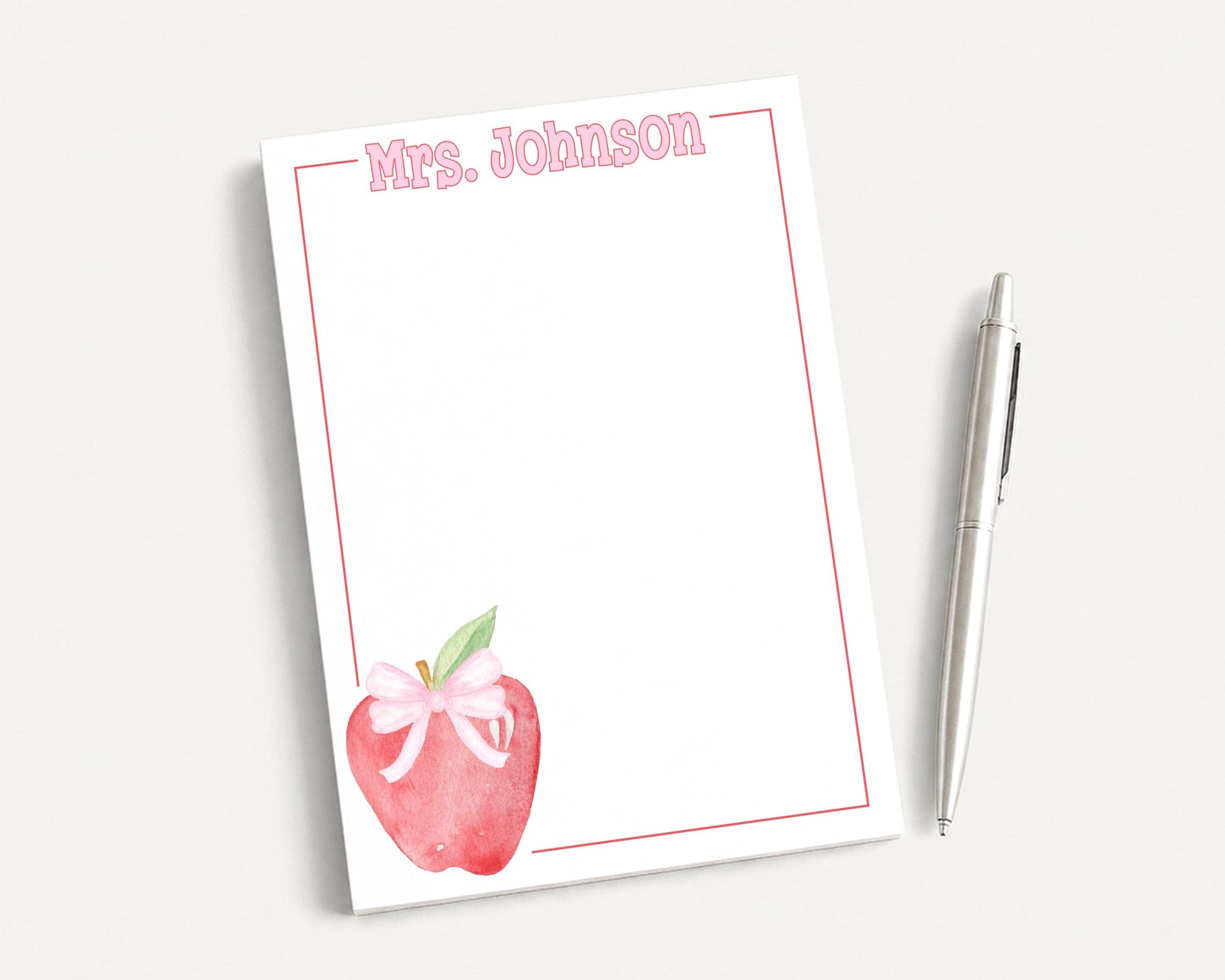 a blank white notepad with a watercolor illustration of an apple on the cover, accompanied by a pen on a white surface.
