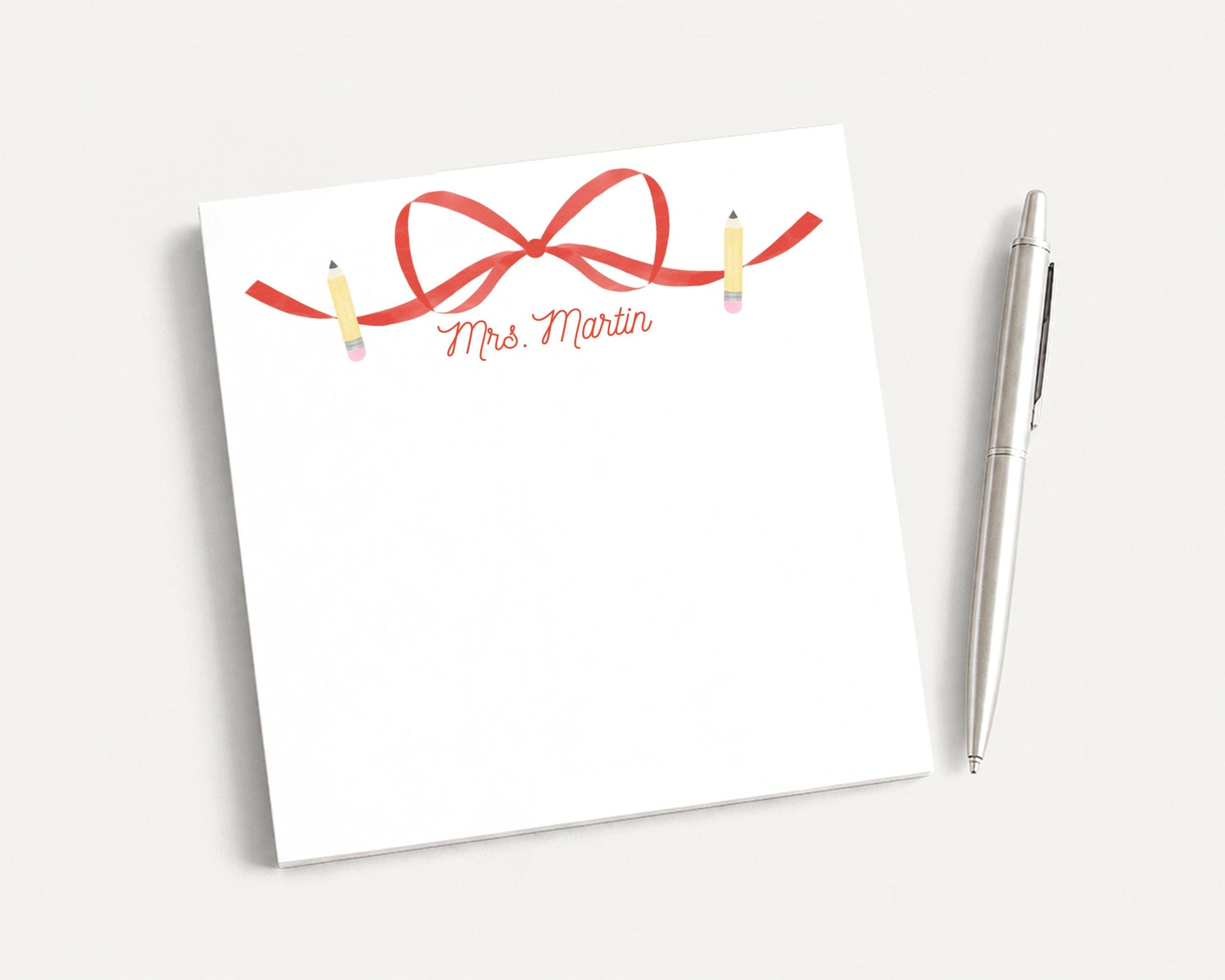 A white notepad with a red ribbon and pencils on it, along with a pen, is placed on a white surface.