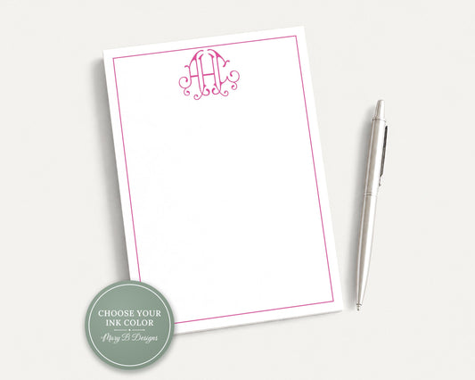 a white notepad with a pink border and a monogrammed logo, accompanied by a silver pen on a plain white background.