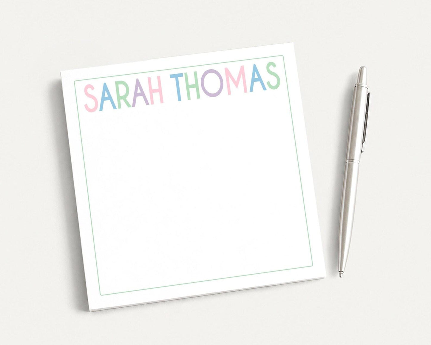A white notepad with the name "SARAH THOMAS" written on it, along with a pen, is placed on a white surface.