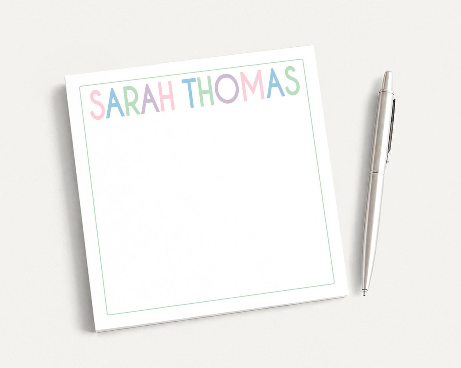 A white notepad with the name "SARAH THOMAS" written on it, along with a pen, is placed on a white surface.