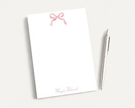 A white notepad with a pink bow and a pen are placed on a white surface.