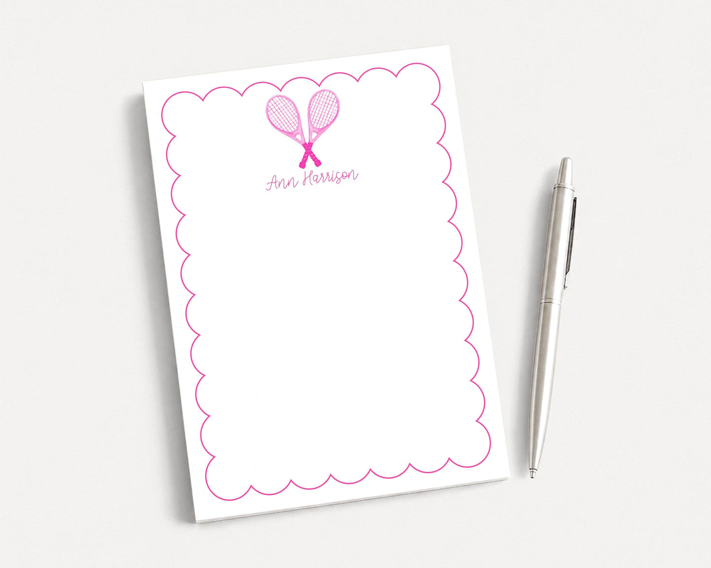 a white notepad with a scalloped edge and a pink tennis racket design, accompanied by a pen on a plain white background.