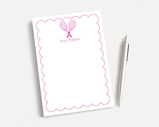 a white notepad with a scalloped edge and a pink tennis racket design, accompanied by a pen on a plain white background.