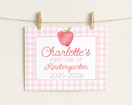 A pink and white checkered sign with a red apple and the text "Charlotte's first day of Kindergarten 2025-2026" attached to it.