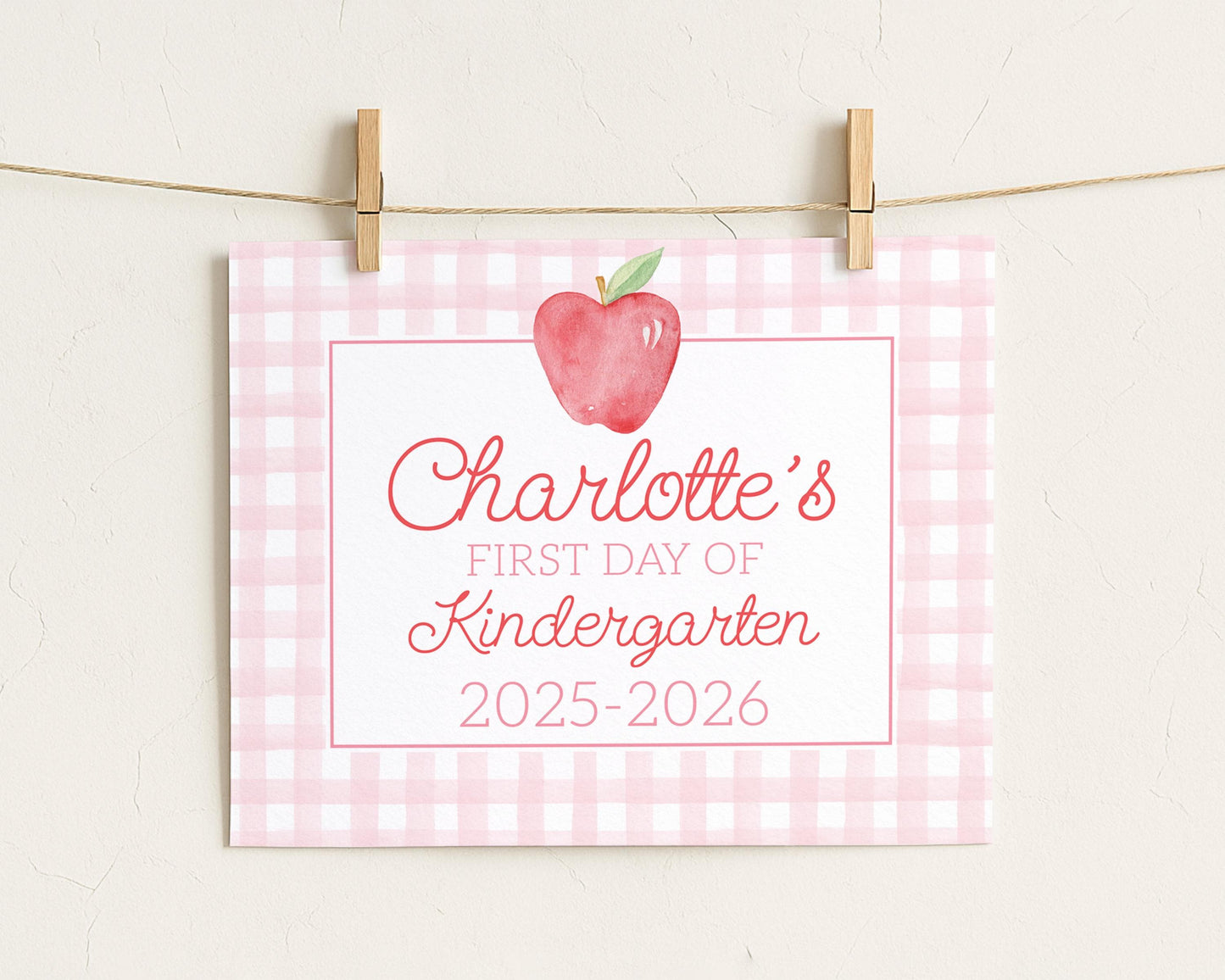 A pink and white checkered sign with a red apple and the text "Charlotte's first day of Kindergarten 2025-2026" attached to it.