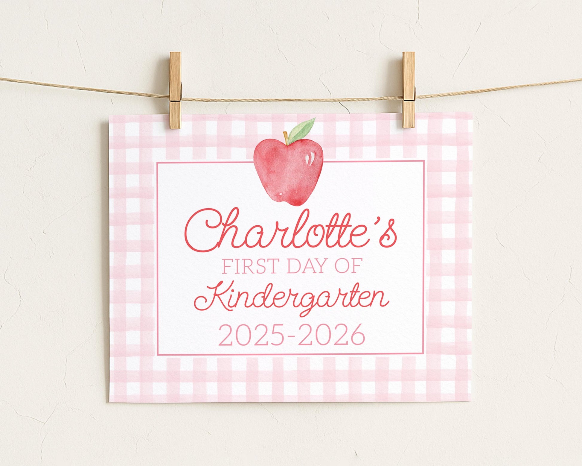 A pink and white checkered sign with a red apple and the text "Charlotte's first day of Kindergarten 2025-2026" attached to it.