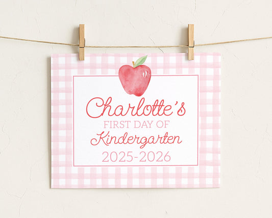 A pink and white checkered sign with a red apple and the text "Charlotte's first day of Kindergarten 2025-2026" attached to it.