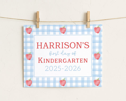 A sign with the text "HARRISON'S first day of Kindergarten" and "2025-2026" printed on it, along with a blue and white checkered pattern and red apples.
