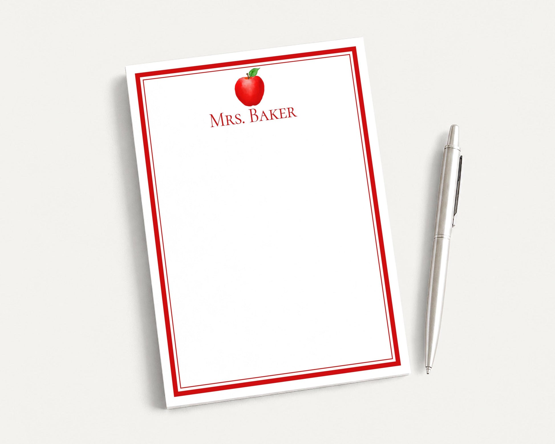 a blank red and white notepad with a red apple design on the top left corner, accompanied by a silver pen on a white surface.