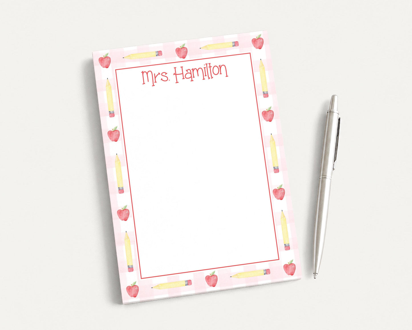 A pink and white notepad with a personalized name and pencil design, accompanied by a pen on a white surface.