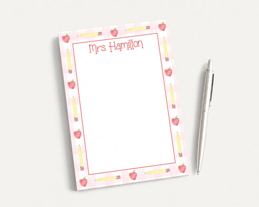 A pink and white notepad with a personalized name and pencil design, accompanied by a pen on a white surface.