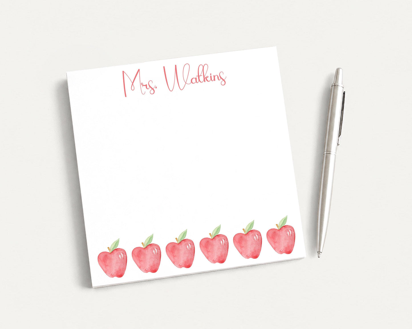 A white notepad with a watercolor illustration of red apples and the name "Mrs. Walker" written on it, along with a pen on a white surface.