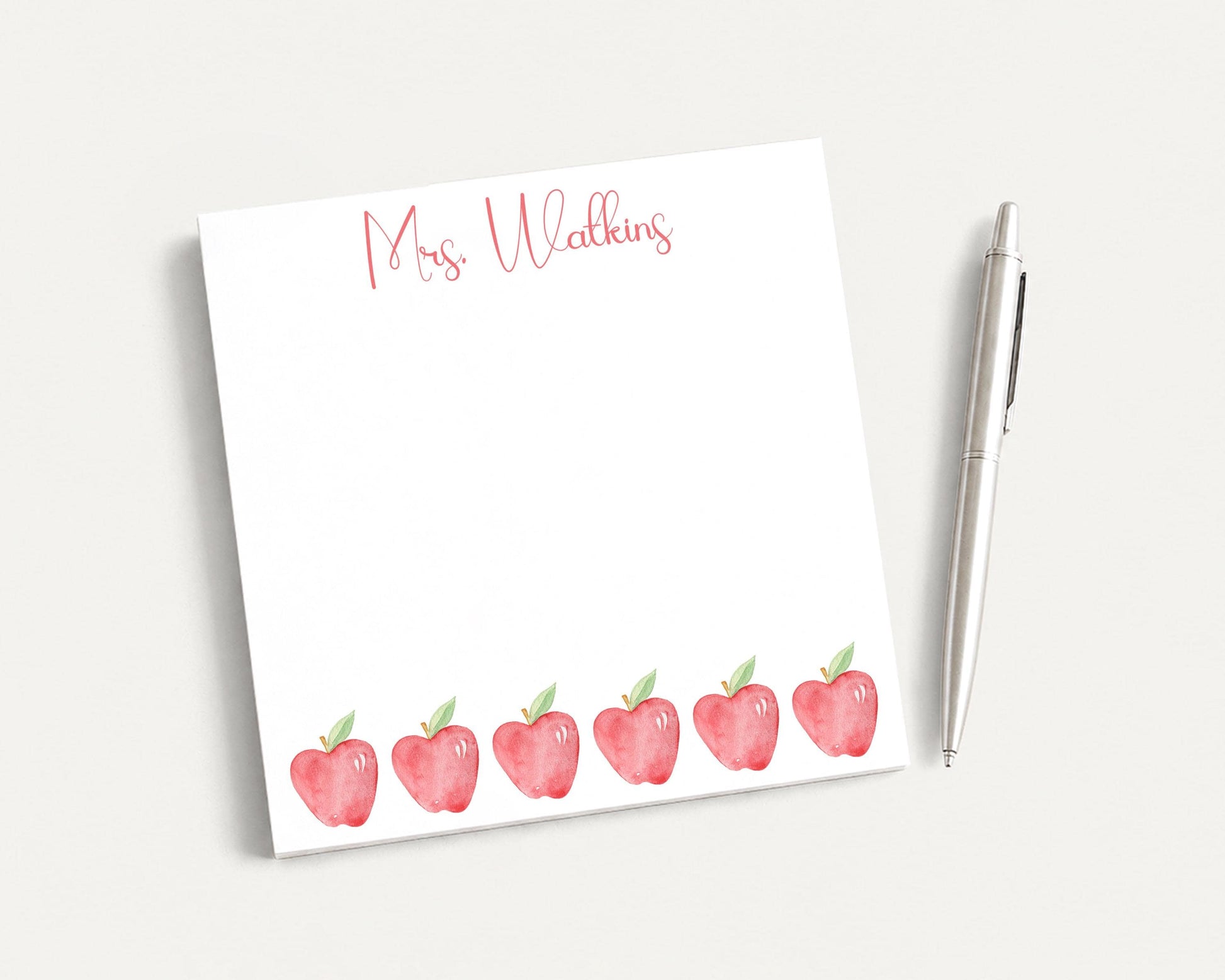 A white notepad with a watercolor illustration of red apples and the name "Mrs. Walker" written on it, along with a pen on a white surface.