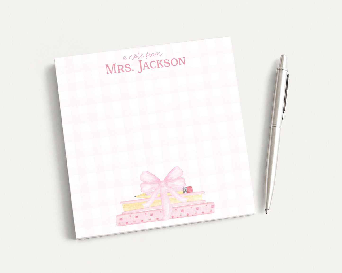A pink and white checkered notepad with a bow on top, accompanied by a pen, on a plain white surface.