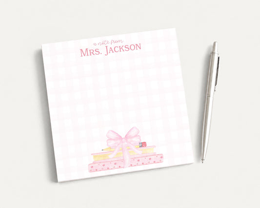 A pink and white checkered notepad with a bow on top, accompanied by a pen, on a plain white surface.