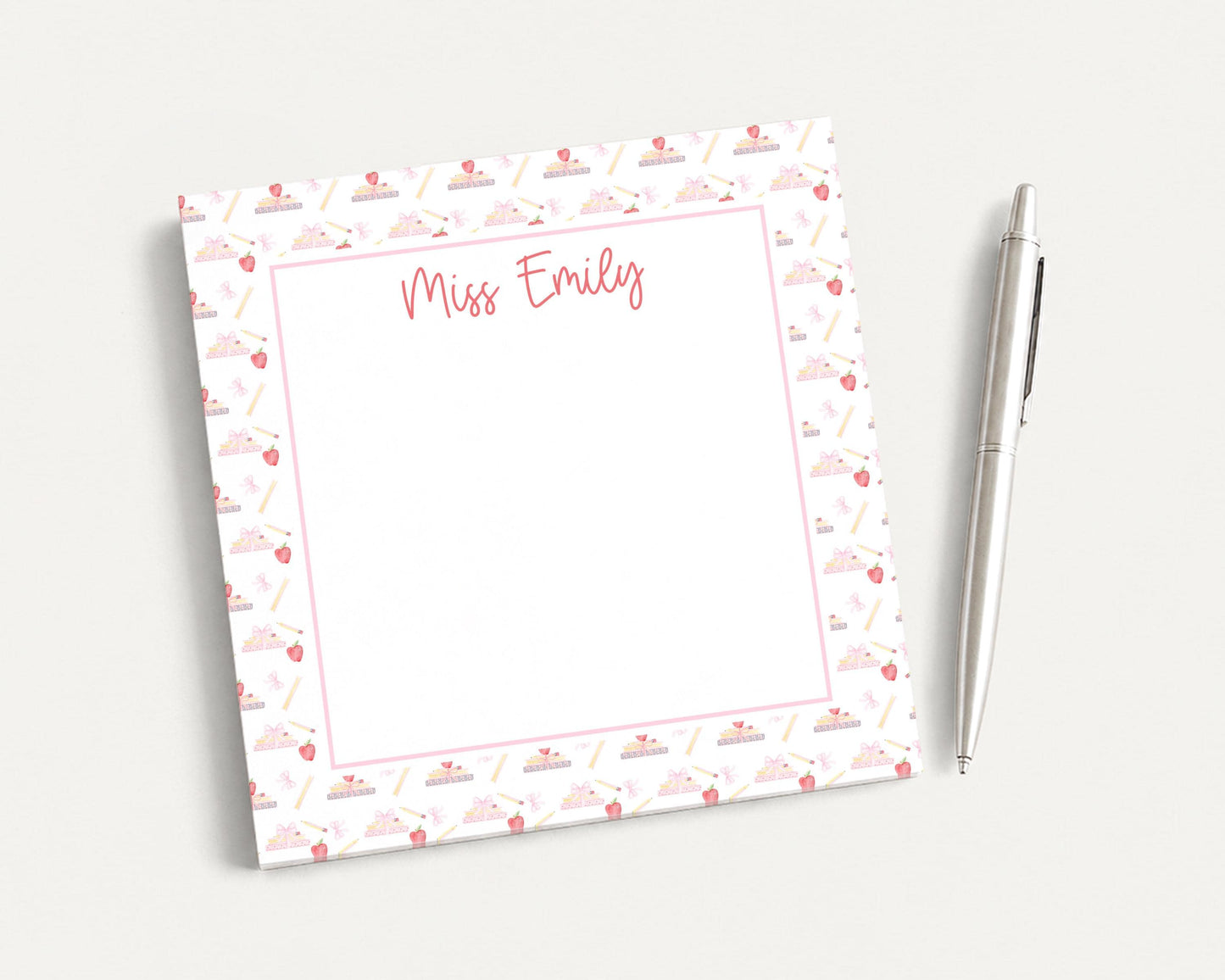 A personalized notepad with a pink and white patterned border, featuring the name "Miss Emily" written in cursive. The notepad is accompanied by a pen, suggesting it is ready for use.