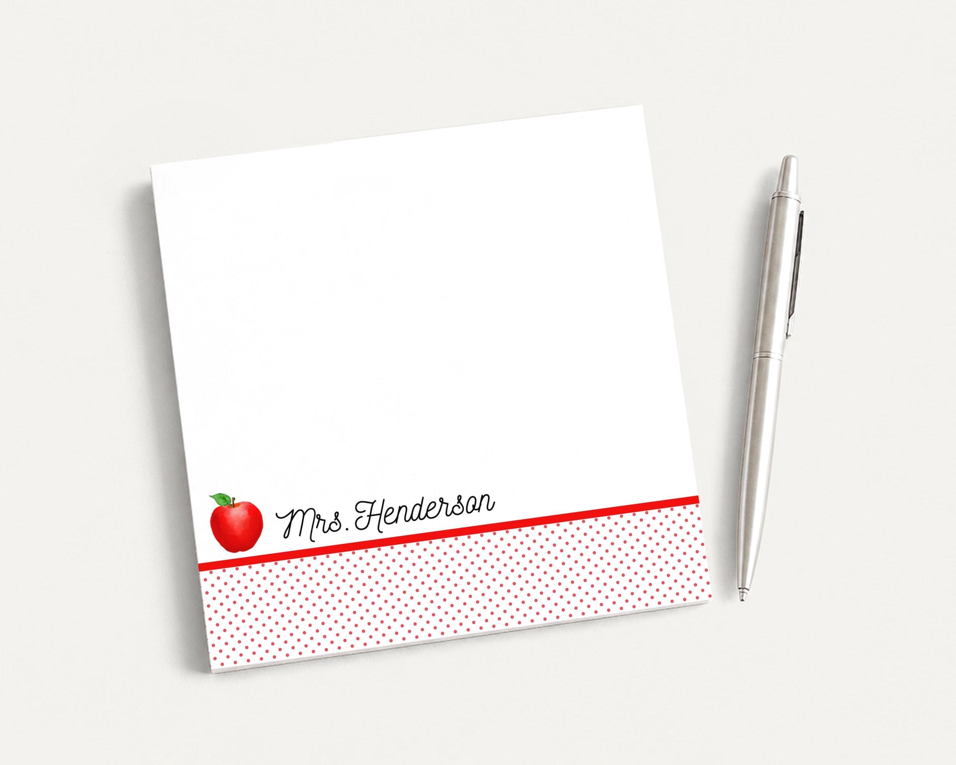 A white notepad with a red border and a red apple graphic, along with a silver pen, are placed on a plain white surface.