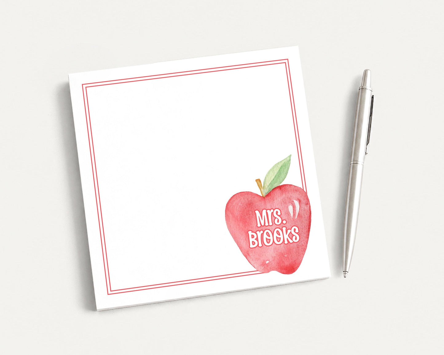 A white notepad with a red border and an apple graphic, along with a pen, is placed on a white surface.