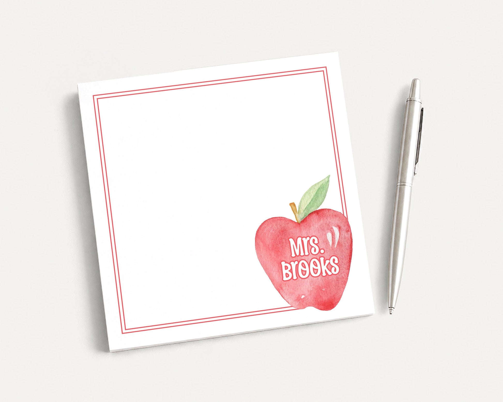 A white notepad with a red border and an apple graphic, along with a pen, is placed on a white surface.