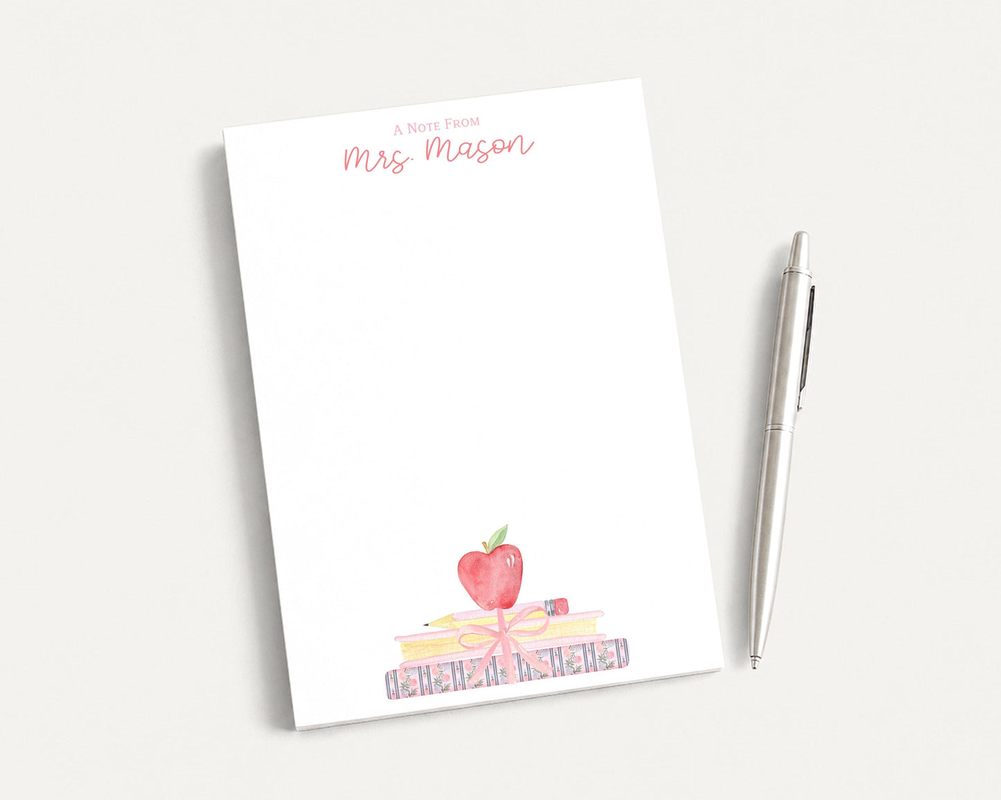 A blank white notepad with a watercolor illustration of a stack of books and an apple on top, accompanied by a pen on a white surface.