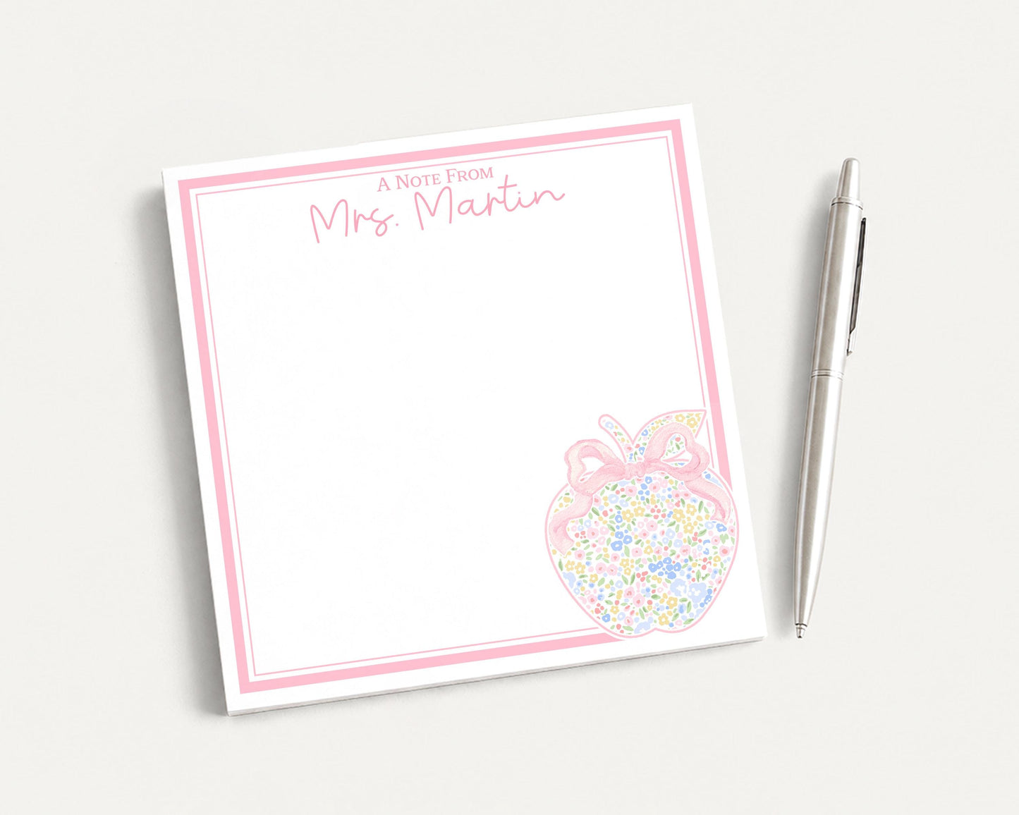 A pink and white note pad with a decorative apple design and a pen on a white surface.