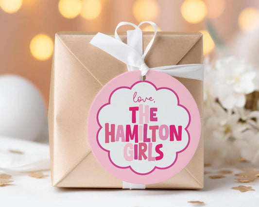 A gift box with a pink and white label that says "love, The Hamilton Girls" is placed on a table with white flowers in the background.