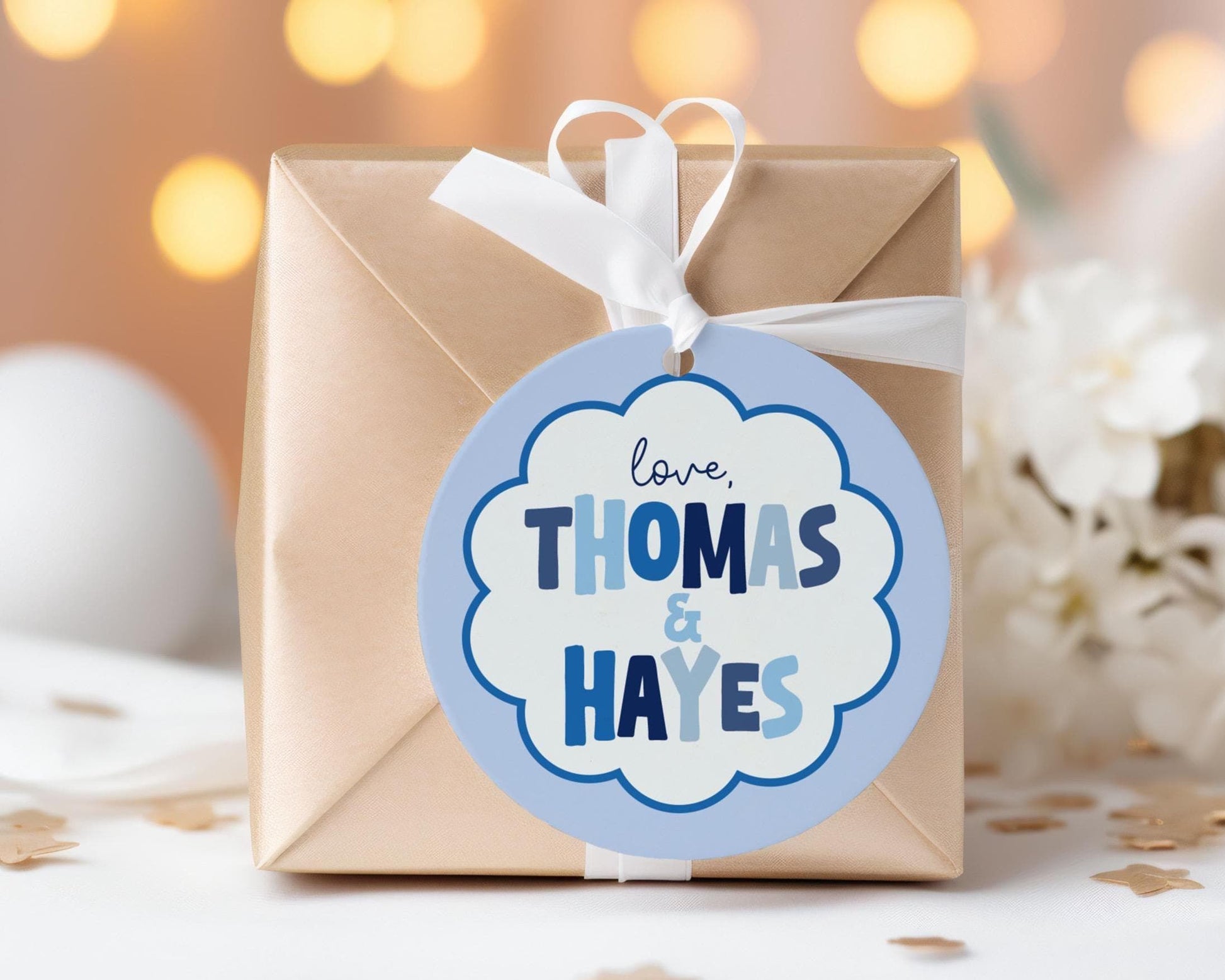 A gift box with a blue and white label that says "love Thomas & Hayes" on it, placed on a table with white flowers and confetti.