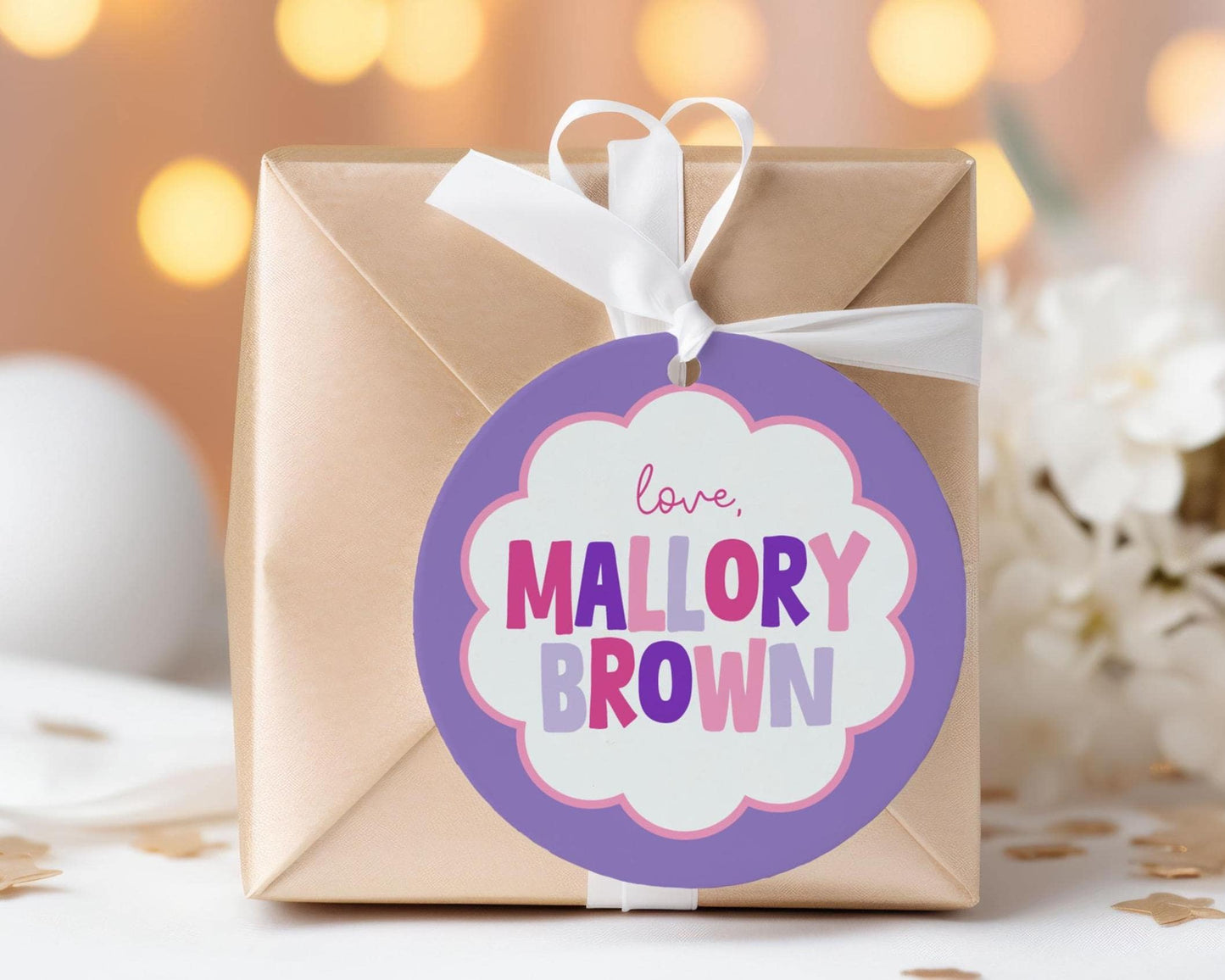 A gift box with a purple and pink label that says "love Mallory Brown" is placed on a table with white flowers in the background.