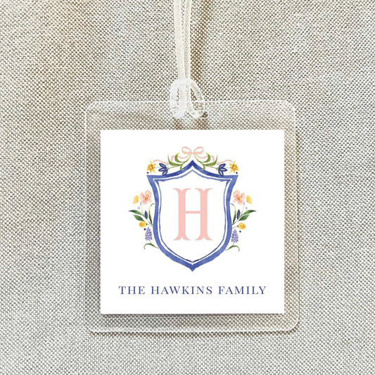 A clear plastic tag with a decorative shield-shaped logo featuring a pink letter "H" surrounded by floral elements. Below the logo, the text "THE HAWKINS FAMILY" is displayed.