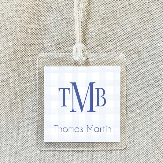 A clear name tag with the initials "TMB" and the name "Thomas Martin" printed on it, hanging on a beige fabric background.
