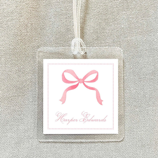 A clear plastic tag with a pink ribbon bow and the name "Harper Edwards" written on it, hanging on a fabric background.