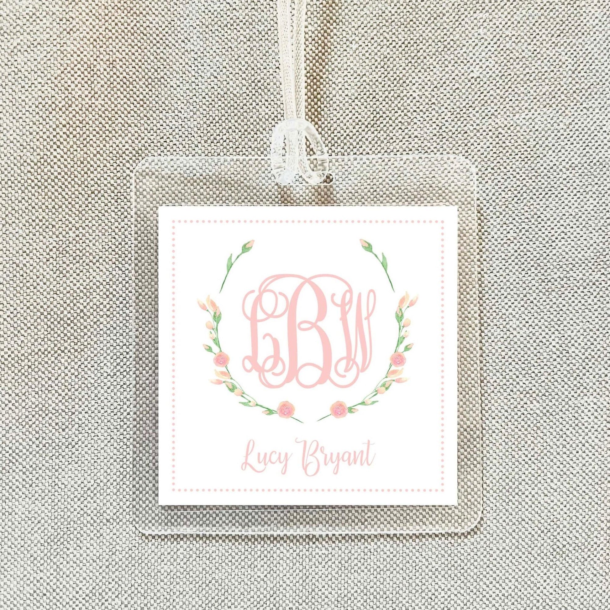 A clear square tag with a monogrammed floral design and the name "Lucy Bryant" printed on it.