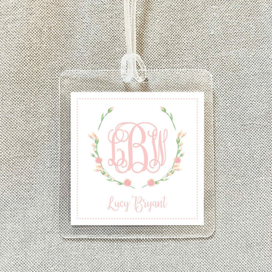 A clear square tag with a monogrammed floral design and the name "Lucy Bryant" printed on it.