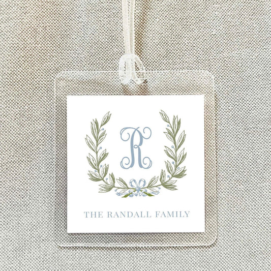 A clear plastic name tag with a monogrammed logo and text, hanging on a beige fabric background.