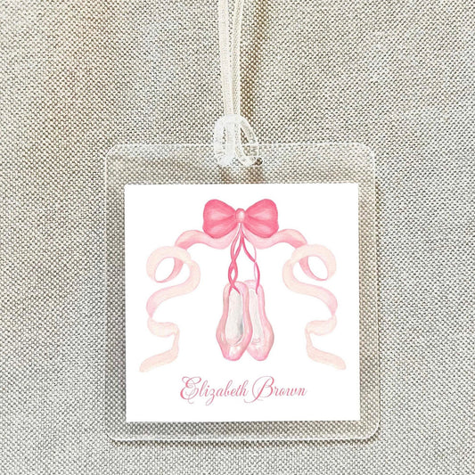 A clear name tag with a pink ribbon and ballet slippers printed on it, hanging on a fabric background.